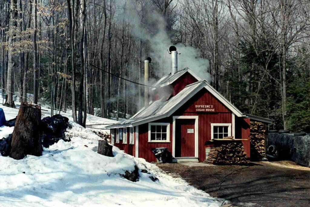 A red barn with multiple stovepips emerging through the roof. The barn is surrounded by snow and has steam billowing from the smokestacks.